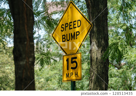 A bright yellow speed bump sign with a 15 MPH limit stands among green trees. The sign is clearly visible, warning drivers in a shaded, wooded neighborhood. A bright yellow speed bump sign with a 15 MPH limit stands among green trees. The sign is clearly visible, warning drivers in a shaded, wooded neighborhood. 128513435