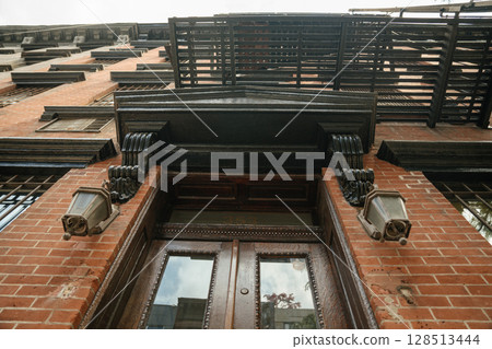 An upward perspective reveals a vintage brick building with ornate black trim, lantern lights, and a metal fire escape. The double wooden doors and classical detailing emphasize historic architecture. 128513444