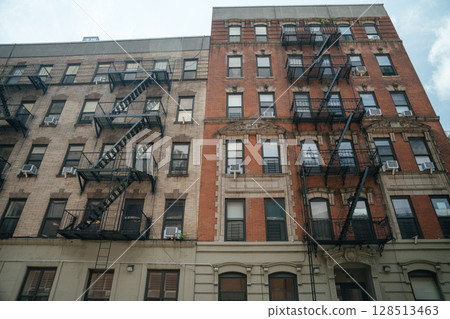 Two classic brick apartment buildings stand side by side, each featuring black metal fire escapes. Window air conditioners and architectural details emphasize the New York City residential style. 128513463