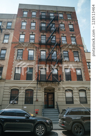 Multi-story brick apartment building features an exterior fire escape, set against a quiet city street. Two black cars parked in front enhance the somber mood of this urban scene during daytime. 128513464