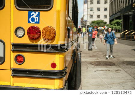 Bright yellow school bus parked on city street features accessibility symbol. Urban environment showcases calm atmosphere under sunny sky, highlighting transportation accessibility. 128513475