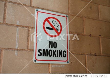 No smoking sign prominently displayed on a brick wall in an outdoor setting. Bright red circle with diagonal line through cigarette conveys cautionary message clearly. No smoking sign prominently displayed on a brick wall in an outdoor setting. Bright red circle with diagonal line through cigarette conveys cautionary message clearly. 128513482