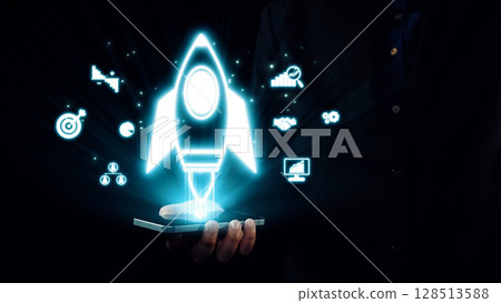 Hand holding smartphone with glowing rocket icon and business-related symbols for innovation, technology, growth, and startup concept in digital environment. Qubit 128513588