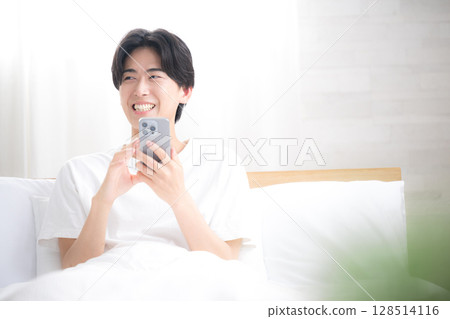 The upper body of a refreshing man relaxing in bed, holding a smartphone and thinking, tapping and looking at the screen 128514116