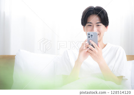 The upper body of a refreshing man relaxing in bed, holding a smartphone and thinking, tapping and looking at the screen 128514120