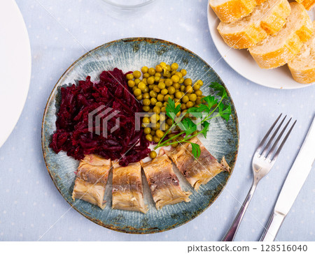 Slices herring fish with canned green peas, beetroot, decorated by mint served on plate with bread and cutlery 128516040