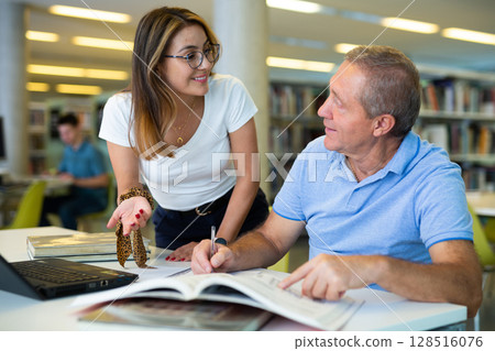 Happy casual Latin woman in glasses talking with older man reading in the library Happy casual Latin woman in glasses talking with older man reading in the library 128516076
