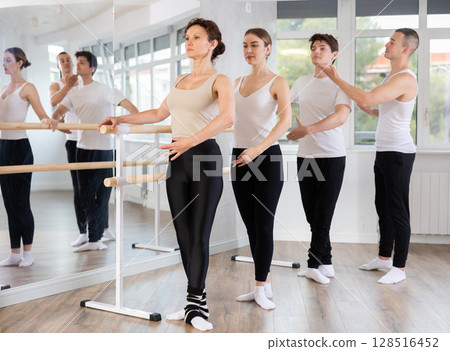 Group of young dancers engaged in ballet in dance studio perfoms exercise near ballet barre, male choreographer helps to coordinate movements correctly 128516452