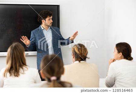 Male business coach standing near interactive board explaining topic while standing against group of people sitting at desks in front of him Male business coach standing near interactive board explaining topic while standing against group of people sitting at desks in front of him 128516463