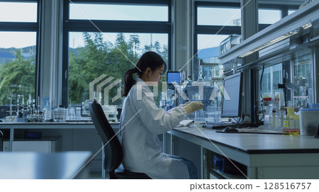 Scientist working modern laboratory with glass shelves filled with scientific equipment and chemicals, wearing white lab coat, focused experiments, bright natural light from large windows 128516757