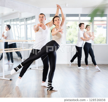 Smiling young woman wearing black and white sportswear dancing in couple and training ballet arabesque pose with partner during group class at dance salon 128516838