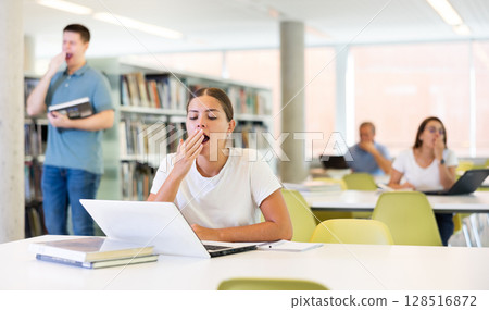 Young unproductive female remote worker yawning while working on laptop computer in the library 128516872