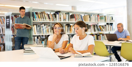 Two friendly adult female students studying in university library, using books and laptop while preparing for lecture Two friendly adult female students studying in university library, using books and laptop while preparing for lecture 128516876