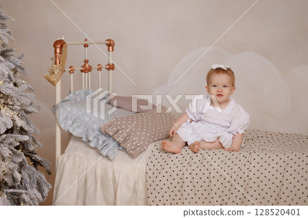 Caucasian baby girl in white dress sitting on bed near Christmas tree in cozy bedroom 128520401