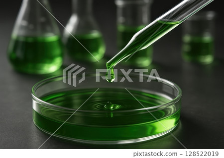 Glass pipette dripping bright green liquid into a petri dish in a laboratory setting 128522019