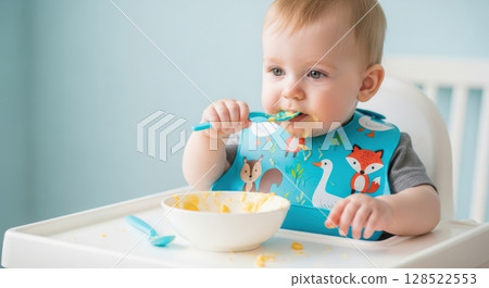Baby in high chair eating pureed food with spoon and bib during mealtime 128522553