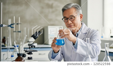Senior asian scientist in lab coat examining blue liquid in flask, conducting research 128522598