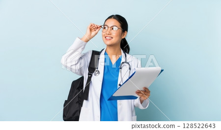 Smiling young asian female medical student in lab coat and scrubs holding clipboard 128522643