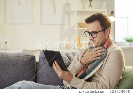Sick young man sitting on sofa at home using tablet with seasonal flu. Sick young man sitting on sofa at home using tablet with seasonal flu. 128524908