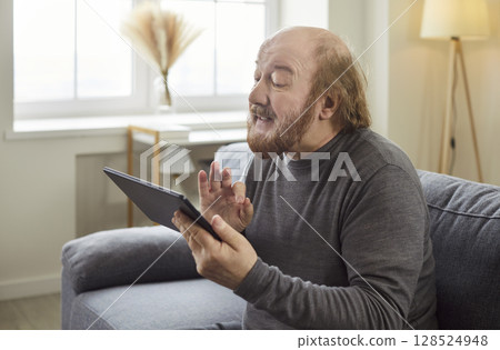 Senior mature man looking at tablet having video call sitting on sofa in living room at home 128524948