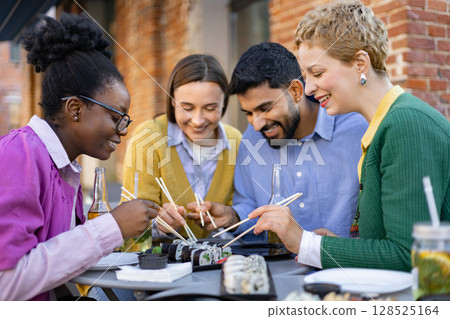 Diverse group of friends enjoying sushi together at an outdoor restaurant, sharing food and laughter. 128525164