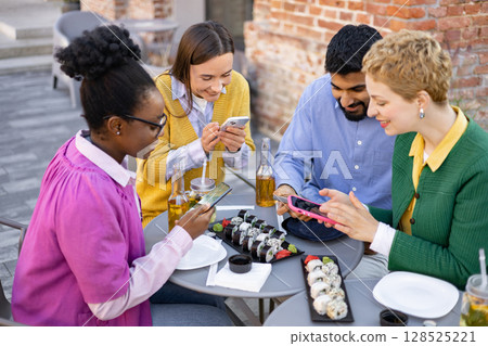A diverse group of friends enjoy sushi and drinks while using their smartphones at an outdoor cafe. 128525221