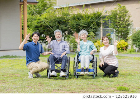Senior couple in wheelchairs waving and their home helper Senior couple in wheelchairs waving and their home helper 128525916
