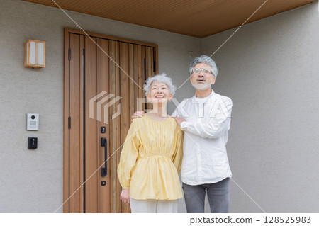 Smiling senior couple standing in front of their house 128525983