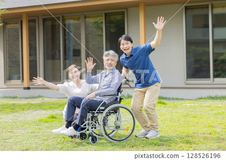 A senior man in a wheelchair and his caregiver waving in front of a house 128526196