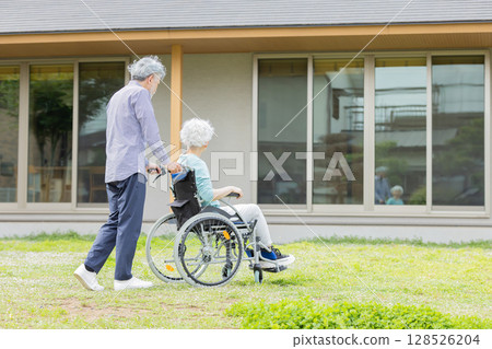 Senior couple in wheelchairs walking in front of their house 128526204