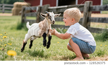 Smiling young boy playing with small goat on sunny farm lawn near wooden fence, showing friendship, childhood joy and rural life. 128526375