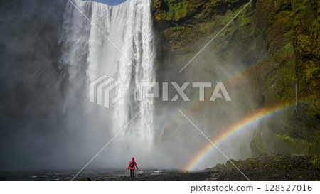 Unidentified woman stands beneath the powerful Skogafoss waterfall in Iceland, framed by mist and a vivid rainbow 128527016