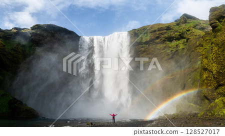 Unidentified woman stands beneath the powerful Skogafoss waterfall in Iceland, framed by mist and a vivid rainbow Unidentified woman stands beneath the powerful Skogafoss waterfall in Iceland, framed by mist and a vivid rainbow 128527017