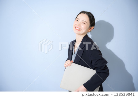 一位年輕可愛的女人穿著西裝站著，手裡拿著筆記型電腦，看著相機，上身，淺藍色背景的商業形象 128529041