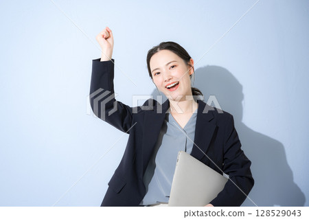 一位穿著西裝、手裡拿著筆記型電腦、握著拳頭等的開朗年輕可愛女子的上半身。淺藍色背景 128529043