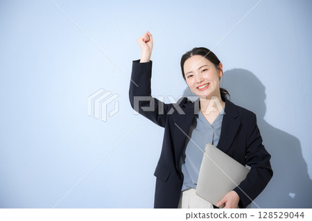 一位穿著西裝、手裡拿著筆記型電腦、握著拳頭等的開朗年輕可愛女子的上半身。淺藍色背景 128529044
