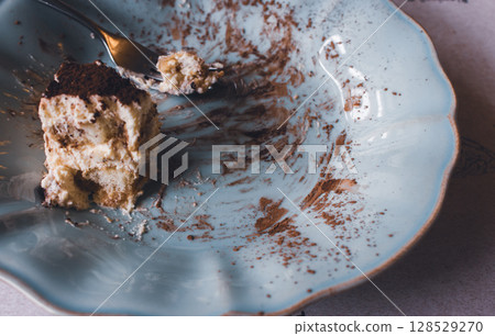 An uneaten piece of cake on blue vintage plate. Slice of tiramisu cake with fork on plate, close up. Delicious chocolate biscuit for breakfast. Sweet food concept. Italian dessert. 128529270