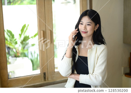 Smiling young woman enjoying a phone call while standing by a bright cafe window Smiling young woman enjoying a phone call while standing by a bright cafe window 128530200