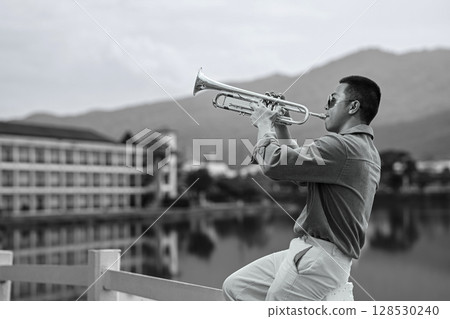 Stylish man performing trumpet on a lakeside balcony with scenic mountain backdrop Stylish man performing trumpet on a lakeside balcony with scenic mountain backdrop 128530240