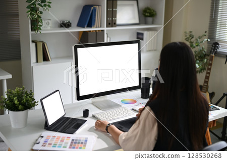 Young female photo editor working on a desktop monitor with camera gear and color charts on the desk 128530268