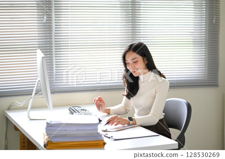 Smiling businesswoman reviewing files at her clean and bright office workstion Smiling businesswoman reviewing files at her clean and bright office workstion 128530269
