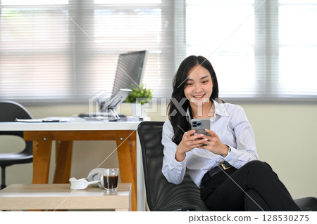Asian office worker enjoys a break relaxing in the office while using her smartphone Asian office worker enjoys a break relaxing in the office while using her smartphone 128530275