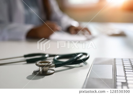 Close up of stethoscope on white desk with doctor writing in the background 128530300