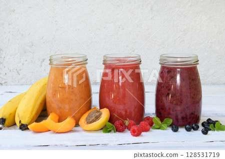 Orange, red and purple smoothies of fresh ingredients - bananas, apricots, raspberries and blueberries on a light background. Healthy food concept. Vegetarian product. Organic Raw Drink. Copy space. Orange, red and purple smoothies of fresh ingredients - bananas, apricots, raspberries and blueberries on a light background. Healthy food concept. Vegetarian product. Organic Raw Drink. Copy space. 128531719