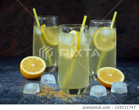 Three glasses of cold homemade lemonade with lemon slices, ice cubes, brown sugar and straws on dark background. 128531747
