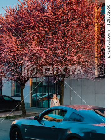 Woman standing calmly on city street behind moving car passing in foreground, vibrant sakura blossoms framing urban spring scenery and creating cinematic depth. Stylish woman standing, car passing 128532050