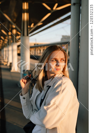 Blonde woman wearing white shirt holding sunglasses standing at railroad platform during golden hour, waiting for approaching train under architectural canopy. Portrait of blonde on train station 128532135