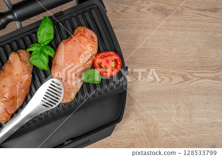 Two raw, marinated chicken breasts with basil leaves and a tomato slice are placed on an electric grill beside a pair of metal tongs on a wooden surface Two raw, marinated chicken breasts with basil leaves and a tomato slice are placed on an electric grill beside a pair of metal tongs on a wooden surface 128533799