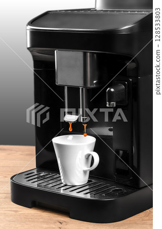 A black modern coffee machine brews espresso into a single white cup placed on a wooden counter with a sleek gradient background 128533803