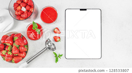 Fresh strawberries, smoothies, and a blender are arranged around a blank tablet screen on a white countertop vibrant kitchen scene. top view. copy space 128533807